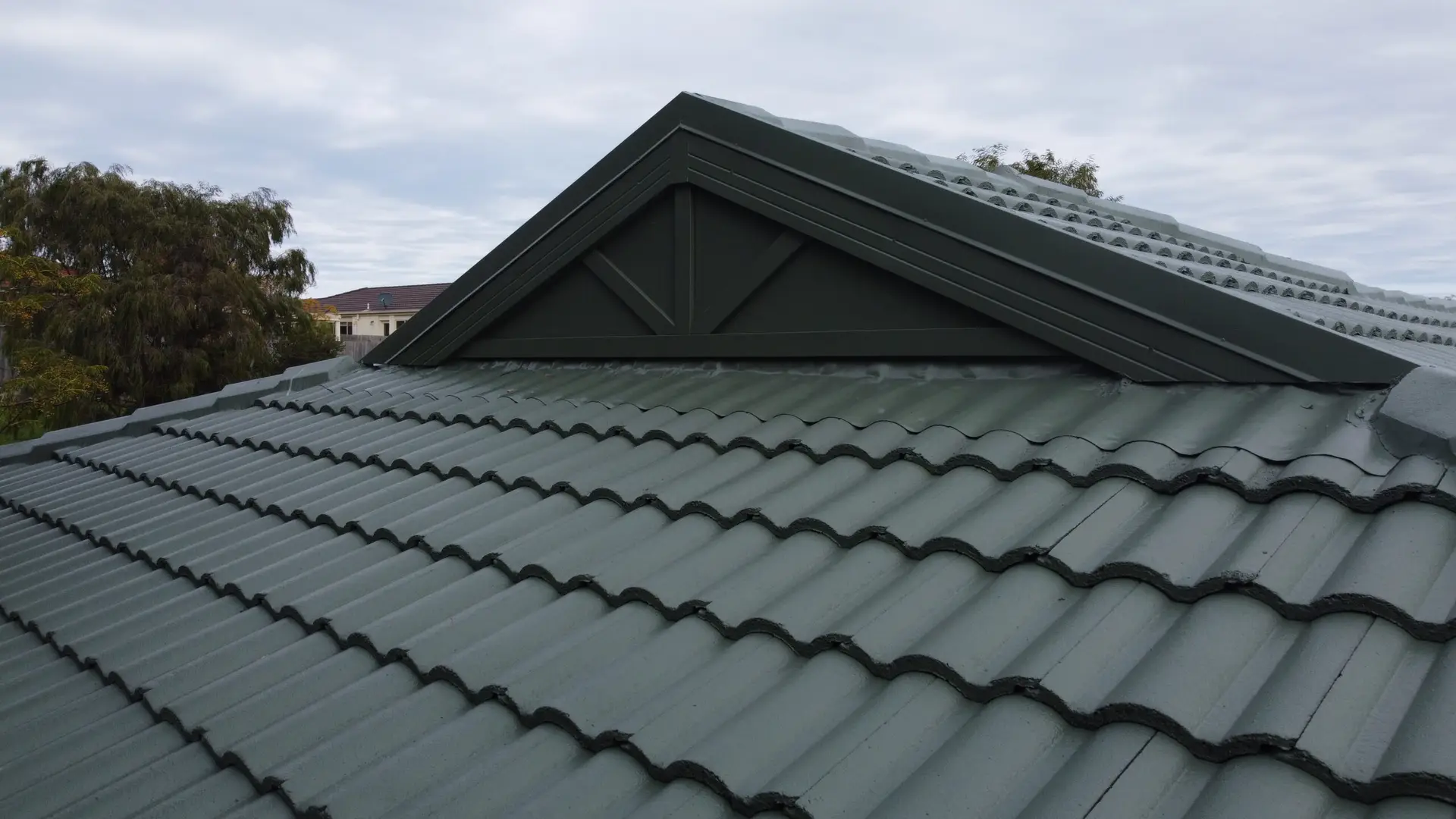 A close-up view of a gabled roof with dark gray, wavy roof tiles, expertly maintained through Tile Roof Restoration Berwick, and a decorative triangular vent beneath the peak. Trees and neighboring rooftops are visible in the background under a cloudy sky.