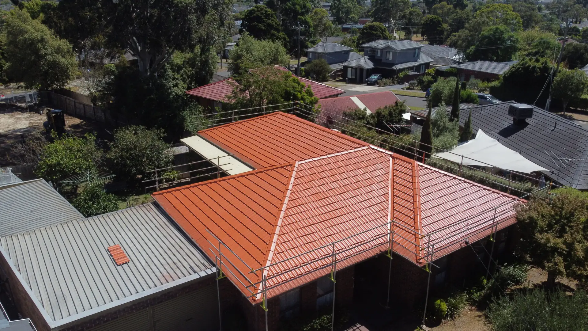Aerial view of a house with a bright red-tiled roof surrounded by trees, nearby homes, and a backyard. The roof is scaffolded, possibly for maintenance or renovation.
