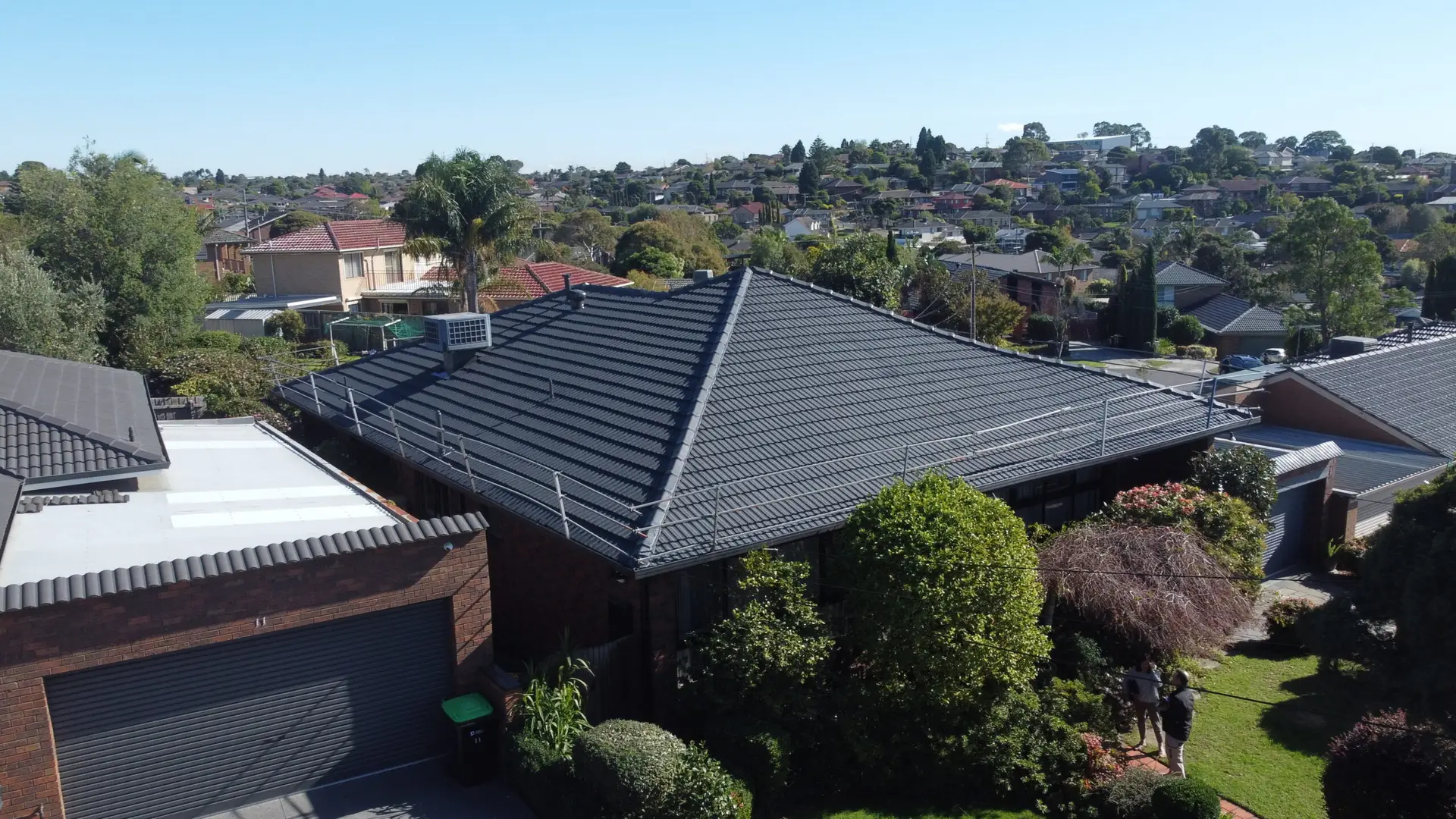 Aerial view of a suburban house with a dark grey tiled roof, surrounded by green trees and bushes, highlighting recent Roof Restoration Dandenong North, with neighboring houses and a clear blue sky in the background.
