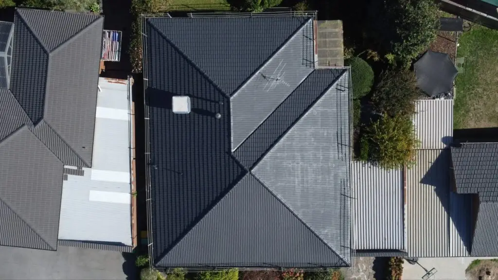 Aerial view of a recently restored dark gray tiled roof completed by Top Glaze Roofing Systems in Melbourne, showcasing precise geometric patterns and a prominent chimney; neighboring homes, driveways, and gardens are visible around the property.