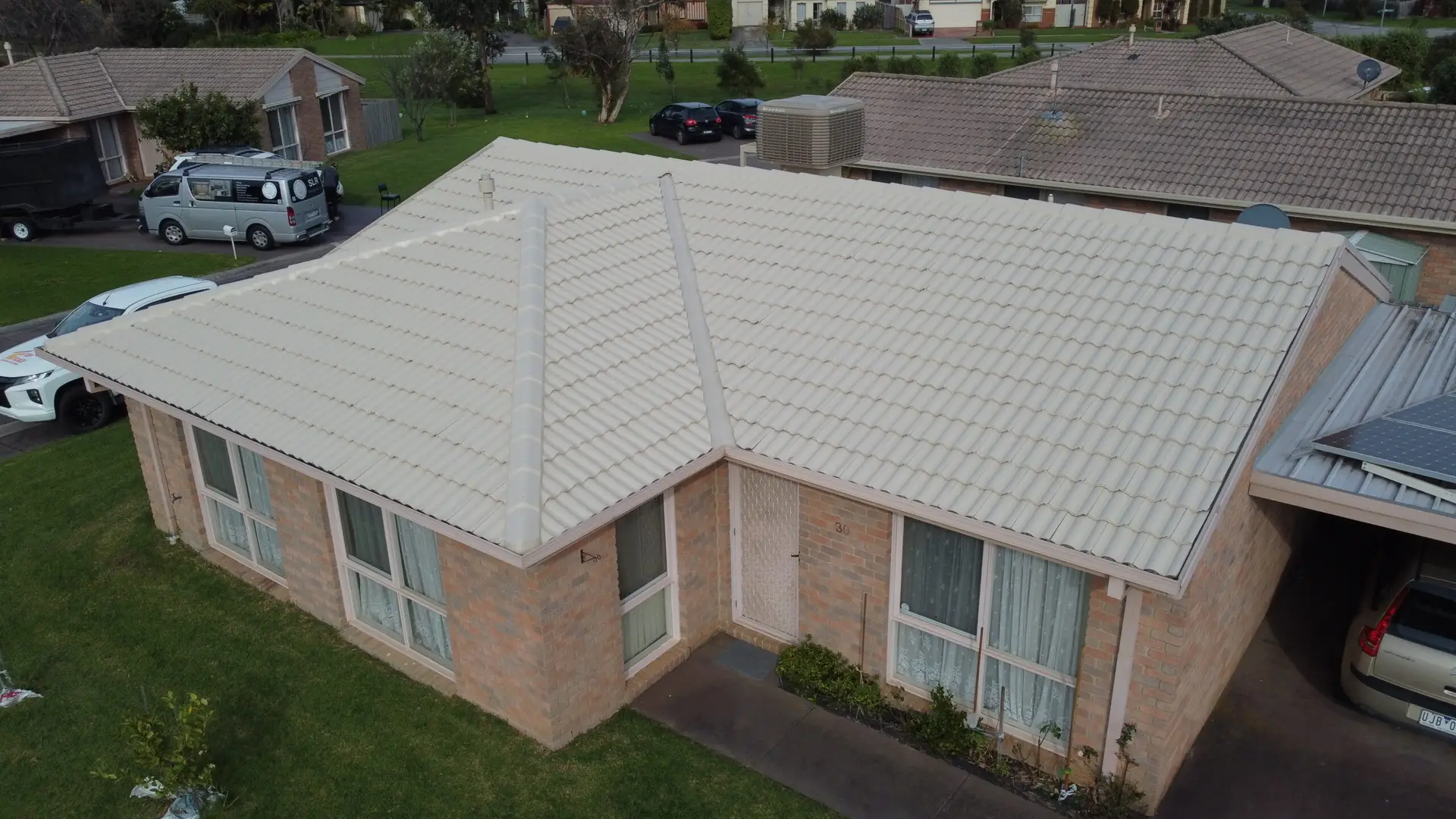 Aerial view of a single-story brick house with a white tiled roof, large windows, a small walkway, and green lawn; neighboring houses and cars are visible in the background—ideal for showcasing Roof Restoration Narre Warren services.