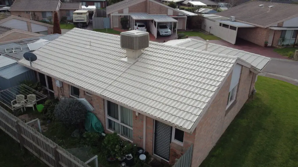 Aerial view of a single-story brick house with a white tiled roof featuring an evaporative cooler. The backyard has a small garden, while neighboring houses and cars are visible—ideal for those considering Roof Restoration Narre Warren.