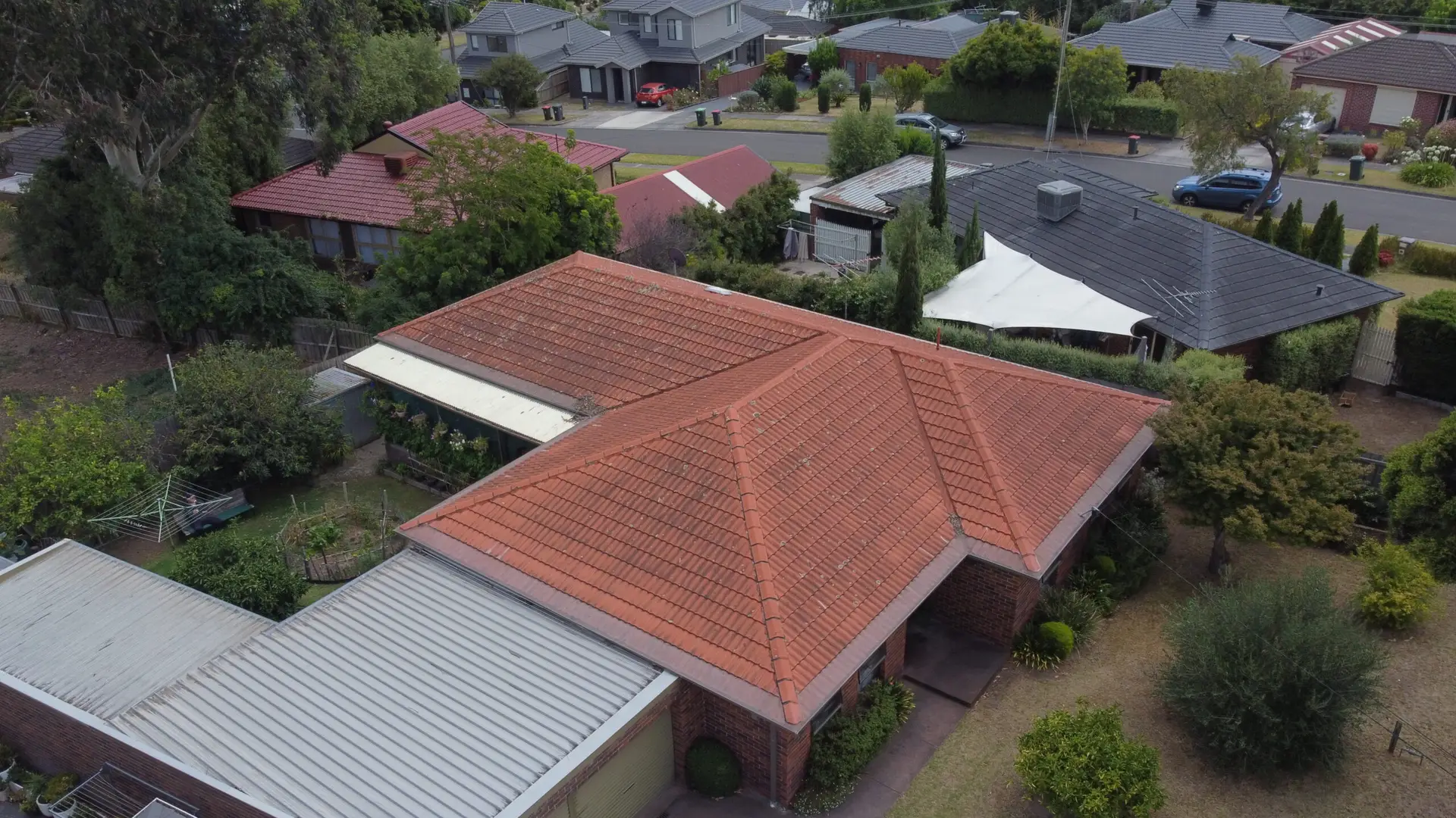 Aerial view of suburban houses with red and grey tiled roofs, surrounded by trees, gardens, and streets in a quiet residential neighborhood.