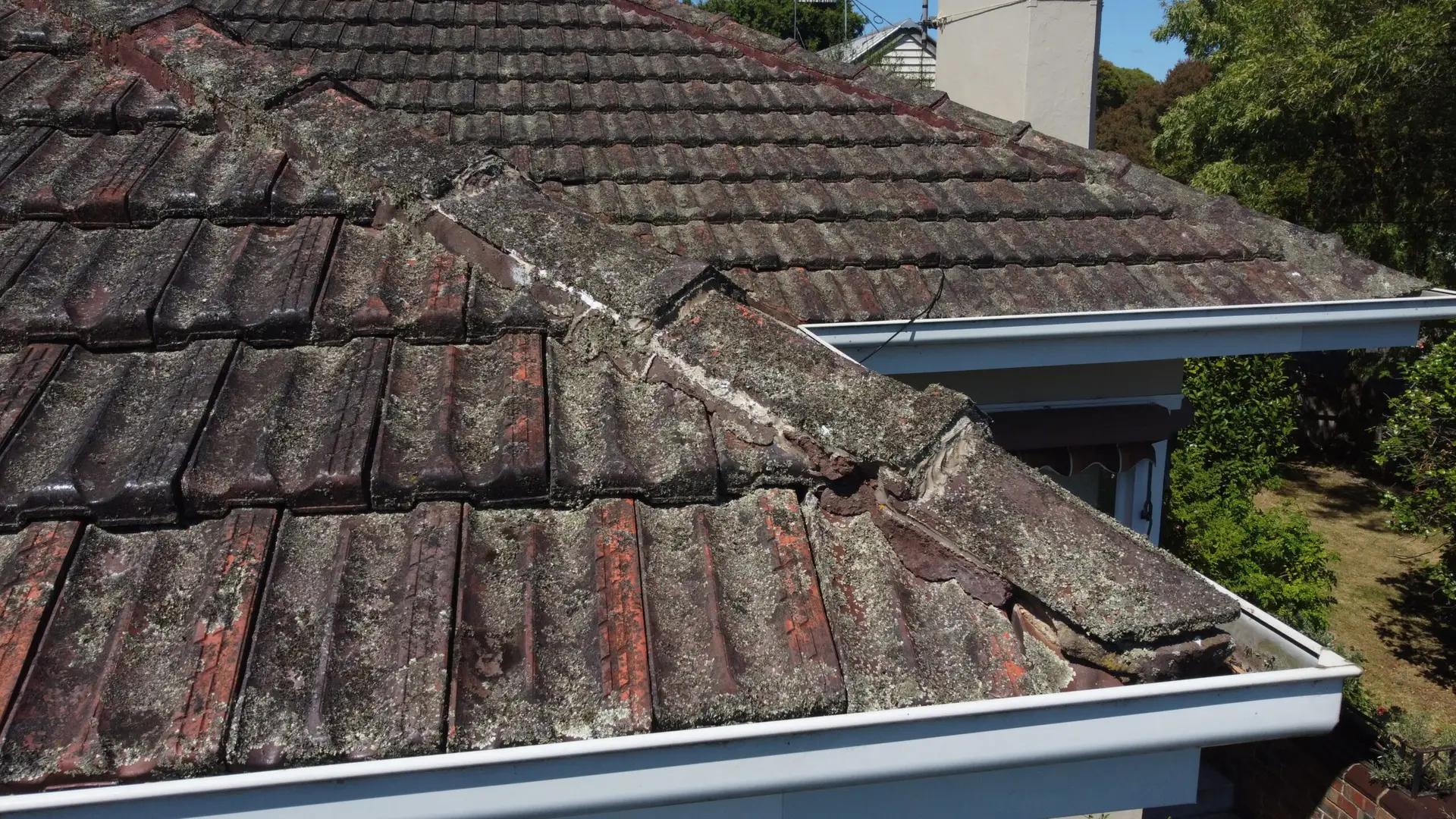 A close-up view of a house roof with old, weathered, and moss-covered tiles. Some tiles are broken and discolored, and the roof appears to need repairs. White gutters run along the edge of the roof.