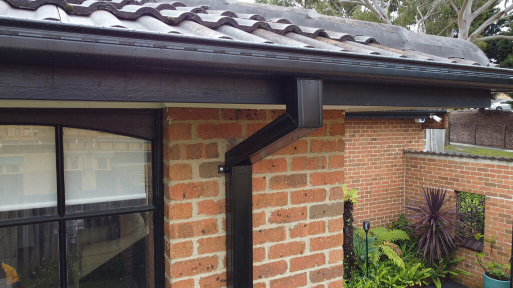 Close-up of a residential roof corner in Melbourne featuring newly installed black gutters and downspout by Top Glaze Roofing Systems, attached to a brick wall with visible roof tiles, window, and surrounding garden plants.