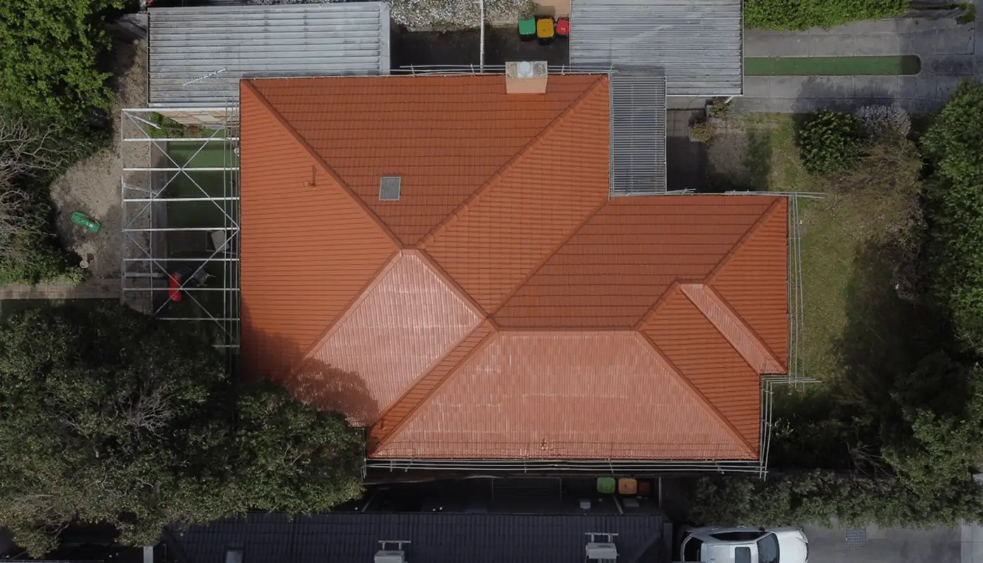 Aerial view of a house with a red-tiled roof, surrounded by trees, adjacent buildings, and parked vehicles, with some green bins visible near the top edge of the image.