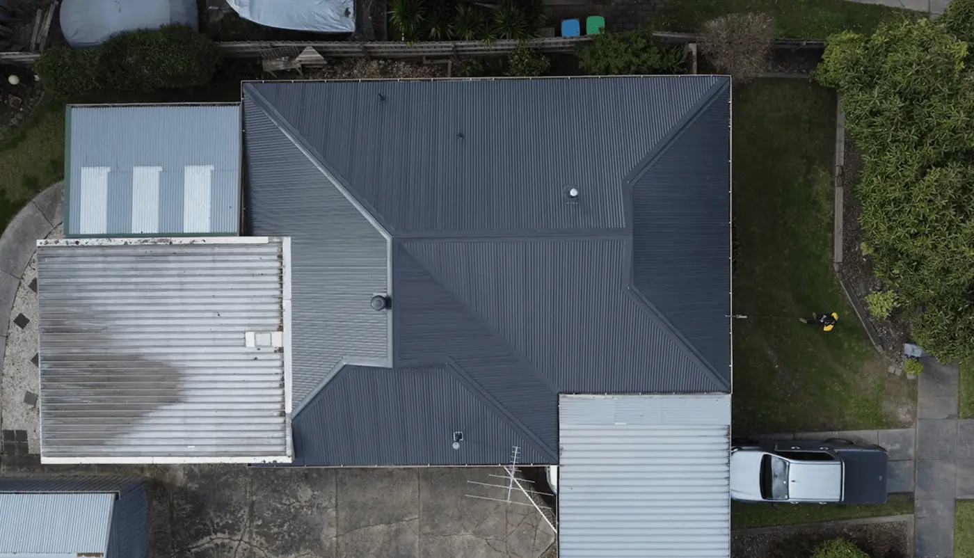 Aerial view of a house with a dark gray roof, surrounded by driveways, green lawns, a white vehicle parked on the right, and various outbuildings and structures around the property.