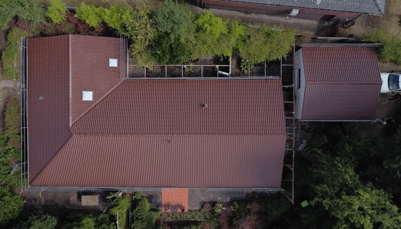 Aerial view of a large house with a red-tiled roof, surrounded by greenery and trees. There are pathways and garden areas visible around the building. Some skylights are present on the roof.
