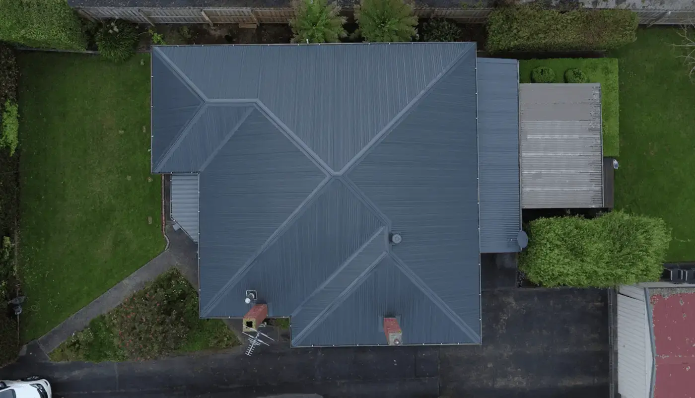 Aerial view of a house with a gray, intersecting gabled metal roof, surrounded by grass, trees, a driveway, and a nearby fenced yard.