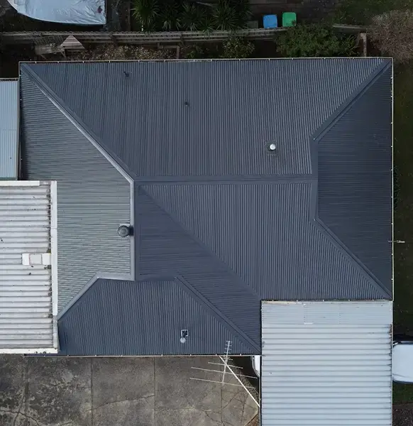 Aerial view of a house roof with dark gray metal panels by Top Glaze Roofing, featuring geometric ridges and a few small vents. Surrounding areas include neighboring fences, greenery, and driveways.