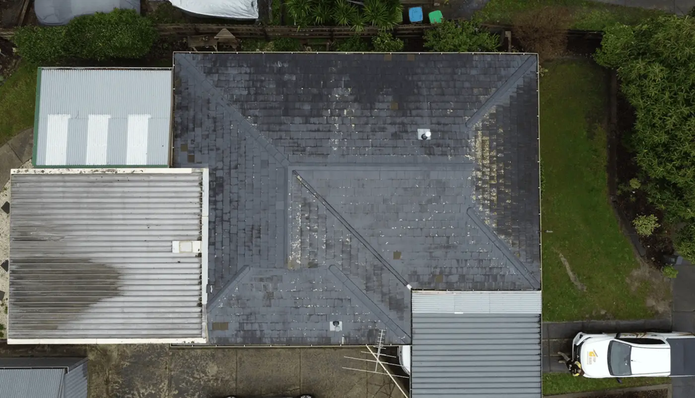Aerial view of a large, dark, weathered roof with visible patches and discoloration, surrounded by smaller metal-roofed structures, greenery, and a white vehicle parked nearby.