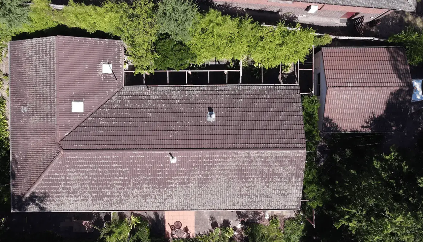 Aerial view of a house with a brown tiled roof surrounded by green trees and plants, with a patio area visible near the bottom and a driveway or road at the top.