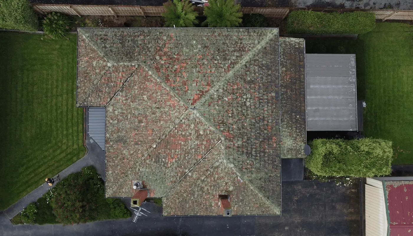 Aerial view of a house roof with a weathered, patchy surface, showing moss and discoloration. Surrounding the house are green lawns, shrubs, driveways, and a wooden fence.