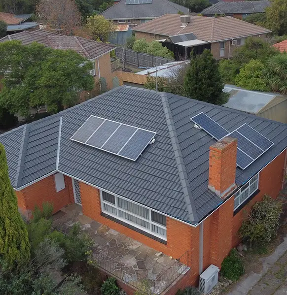 Aerial view of a brick house with a tiled roof by Top Glaze Roofing featuring solar panels. The house is surrounded by trees and neighboring homes in a suburban area.
