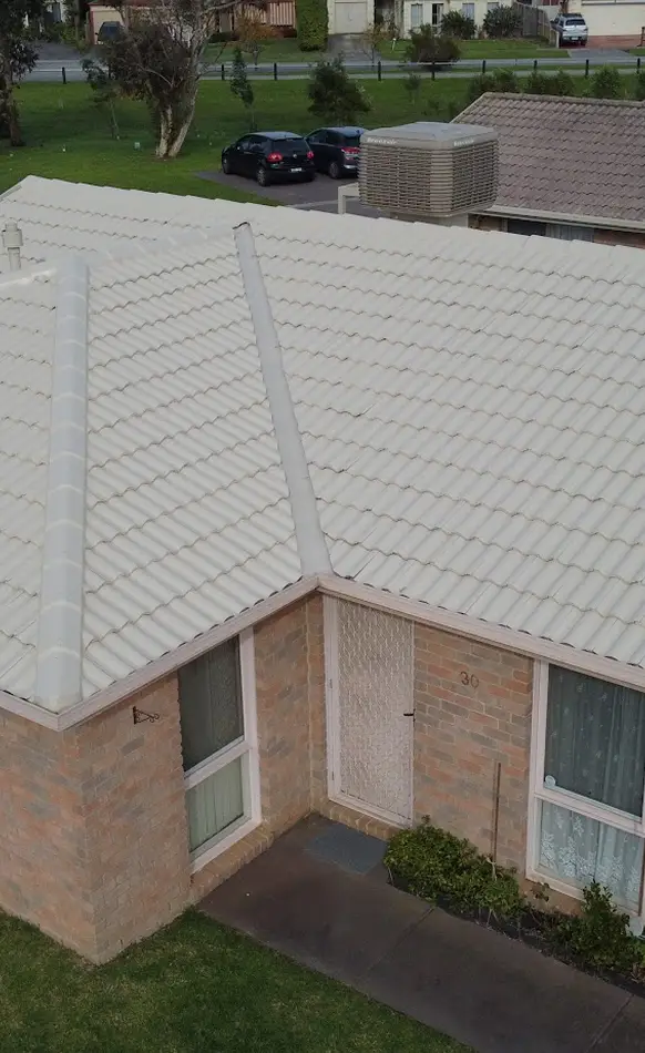 Aerial view of a house with light-colored tiled roof, brick walls, a mesh-covered front door, two windows, and a small garden. Nearby, two cars are parked on a street with grass and trees.