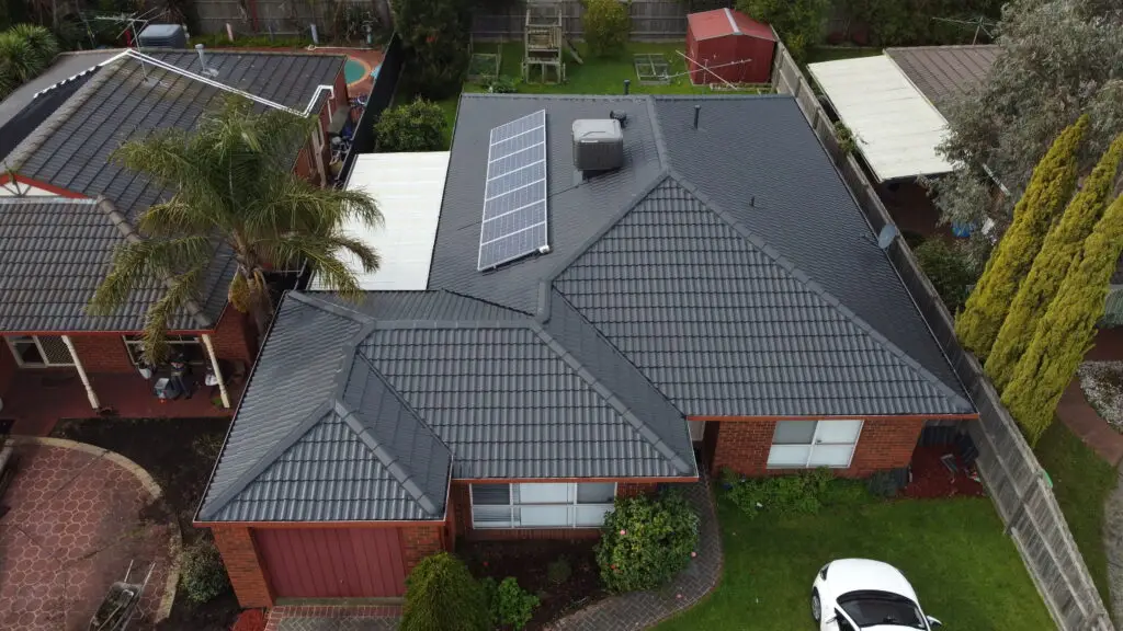 Aerial view of a single-story brick house with a dark tiled roof, solar panels, skylight, and garage. The property features a lush garden and trees. Recent tile roof replacement enhances its appeal. A white car is parked in the driveway.