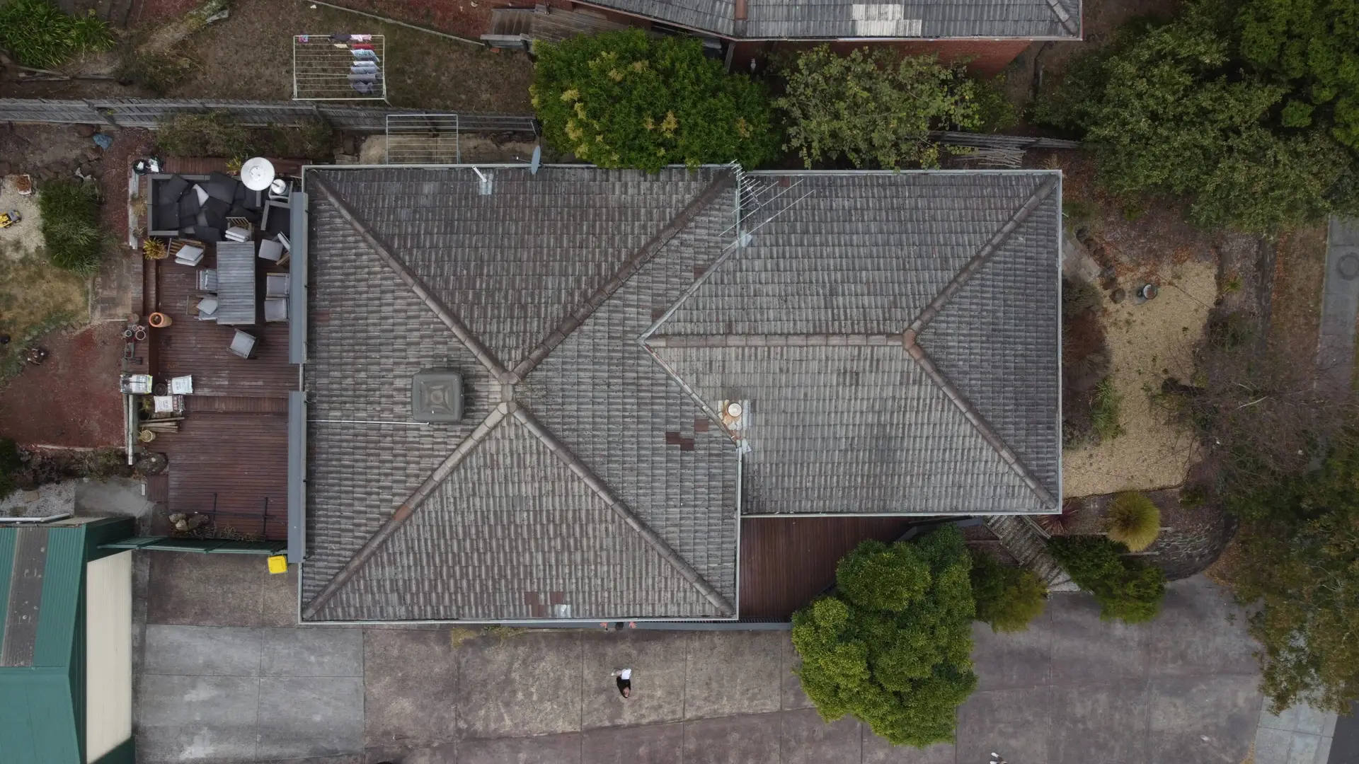 Aerial view of a gray tiled-roof house surrounded by trees, a driveway, and a small backyard area with outdoor furniture and miscellaneous items. The house has a symmetrical, cross-shaped roof design.