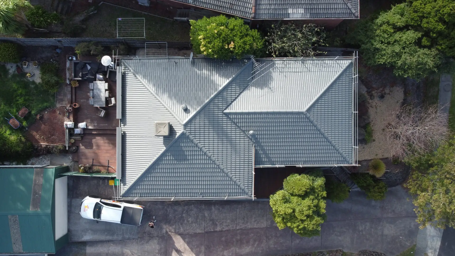 Aerial view of a house with a grey, gable-style roof, surrounded by trees. A white vehicle is parked in the driveway, and a deck area with outdoor furniture is visible on the left side of the house.