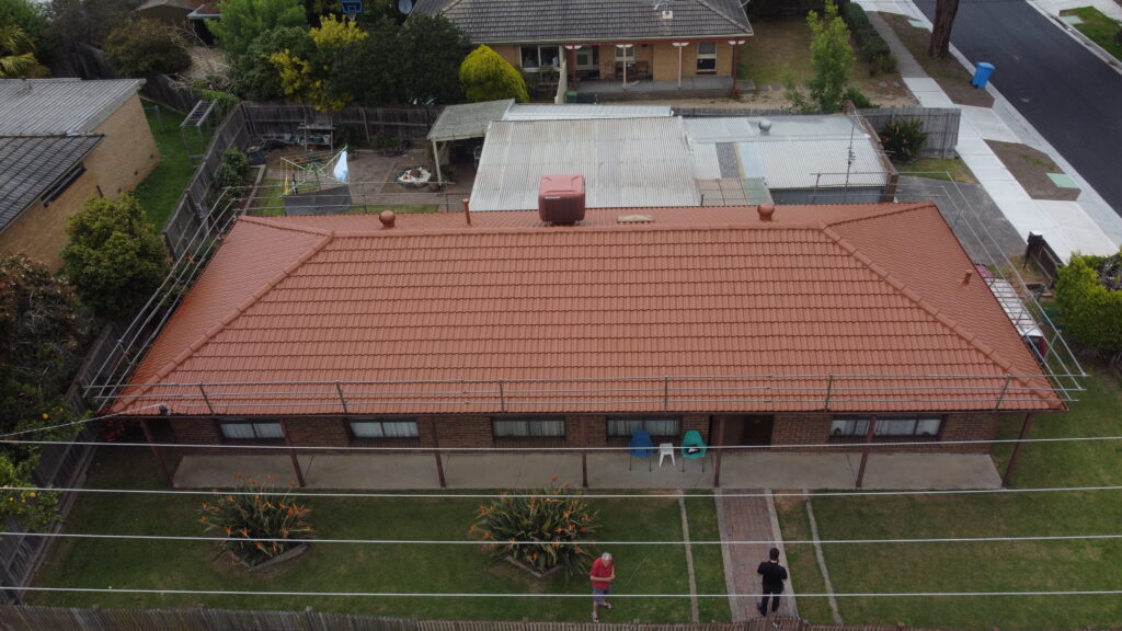 Angled aerial shot of a Cranbourne brick house featuring a freshly restored red tiled roof, surrounded by scaffolding, trees, and neighbouring rooftops.