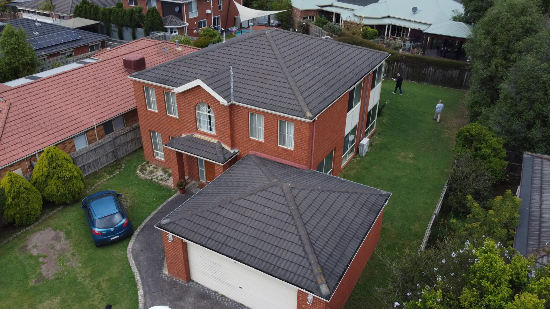 Aerial view of a large two-story brick house with a gray tiled roof, a driveway with a blue car, a front yard with bushes, and three people standing in the backyard.`