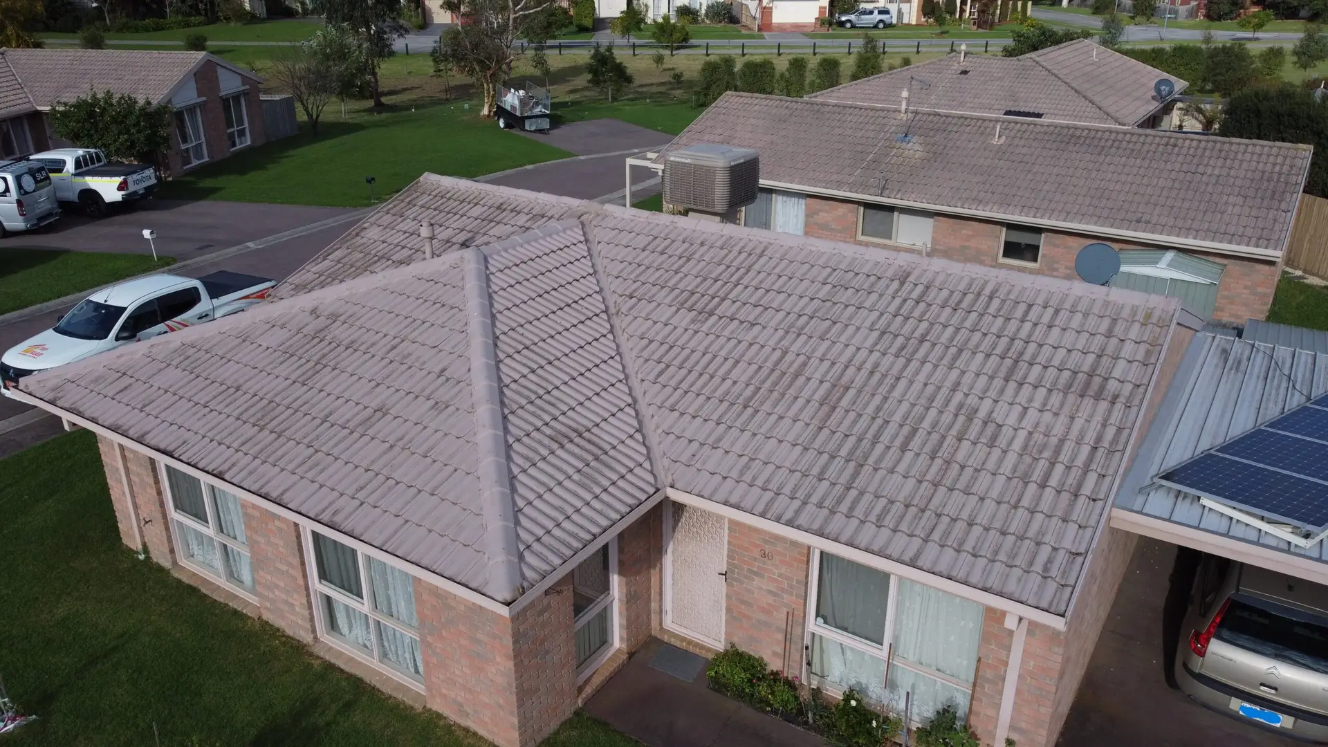 Aerial view of a single-story brick house with a tiled roof, large windows, and a small garden. Another similar house and grassy areas are visible in the background.