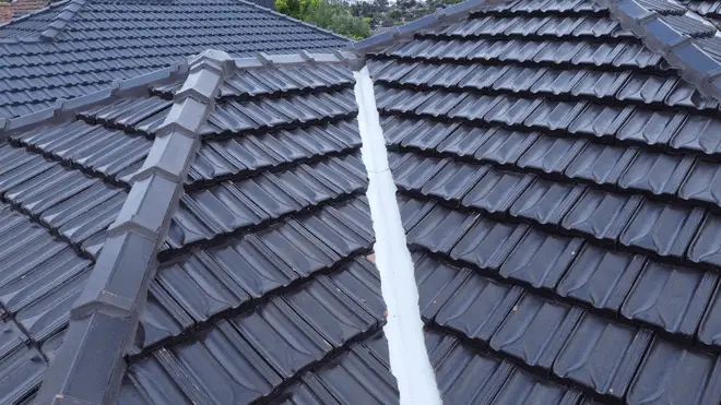 Close-up view of a tiled roof with dark grey ceramic tiles and a central white sealant strip along the roof valley. The detailed location also reveals other roof sections and trees visible in the background.