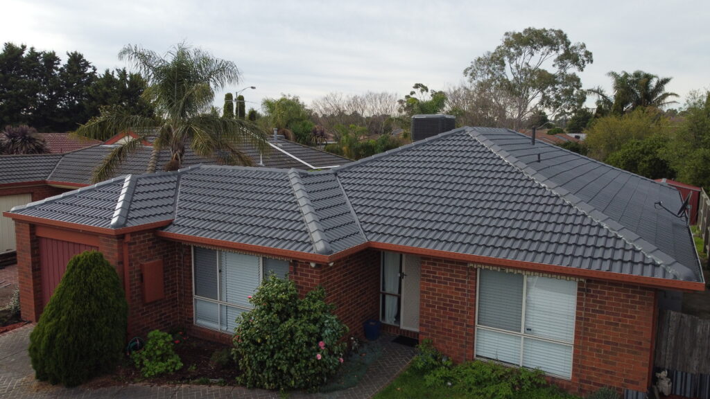 Close-up aerial view of a charcoal grey tile roof in Cranbourne with vent pipes and neatly sealed flashing, surrounded by garden beds, fencing, and light green foliage.