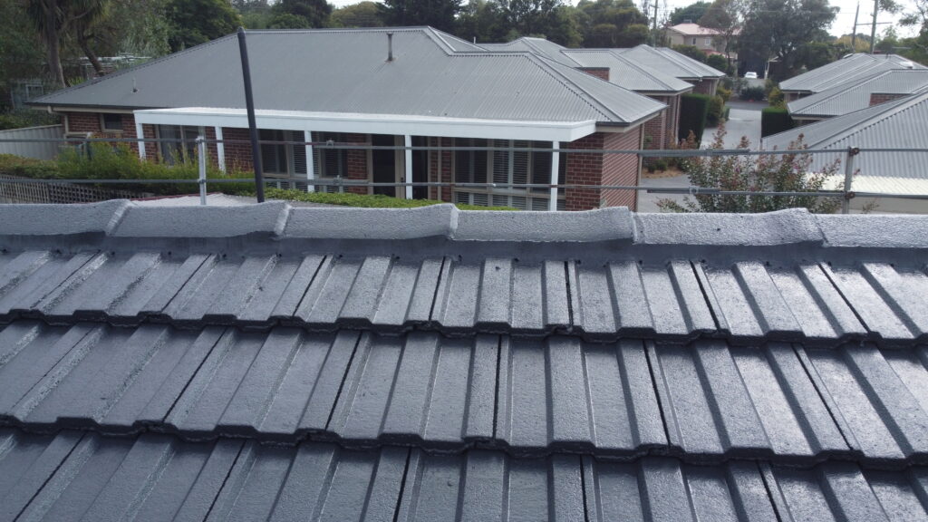 Close-up of a restored dark grey tile roof in Croydon with fresh ridge capping, overlooking a row of brick homes with light grey metal roofs and leafy surroundings.